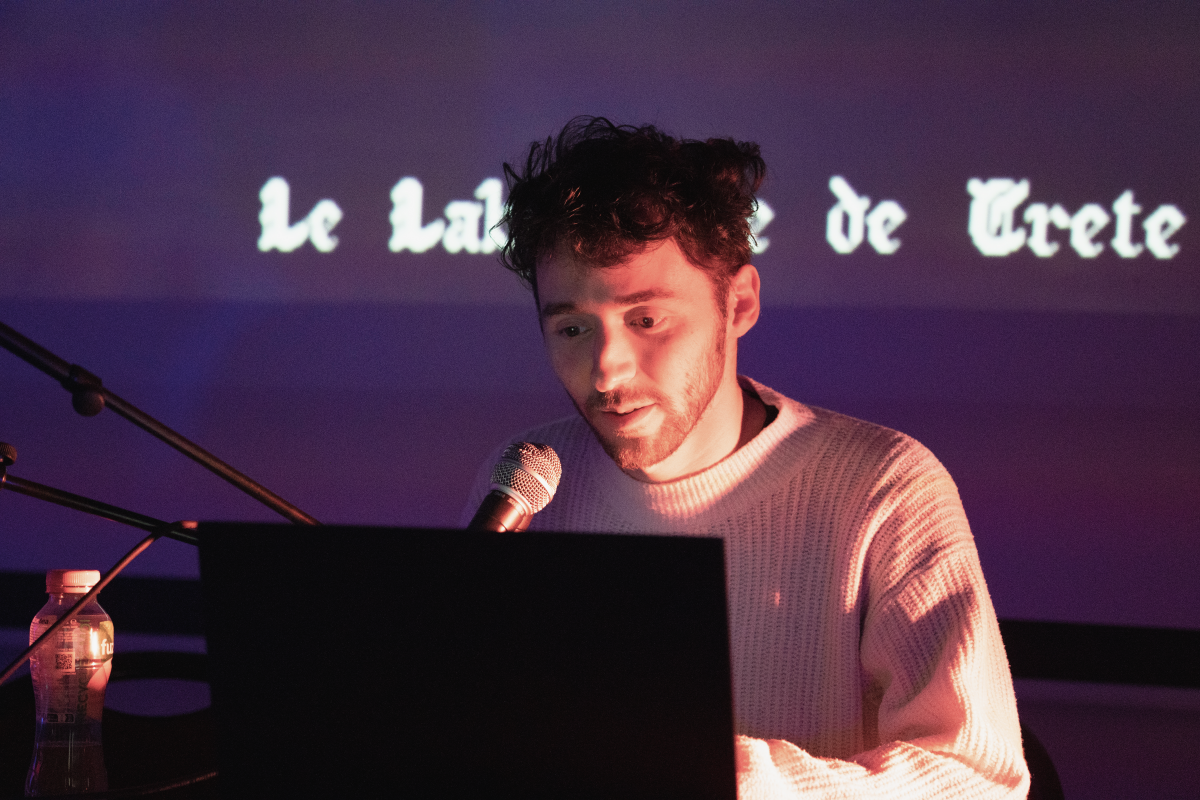 Person speaking into a microphone while reading from a laptop, with projected text 'Le Labyrinthe de Crete' in the background, under warm stage lighting.