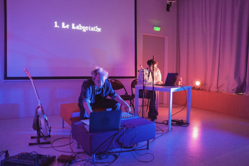 Musician playing electronic keyboard and person reading from a laptop in a presentation room with text 'Le Labyrinthe' projected on a screen, ambient lighting setting a calm atmosphere.