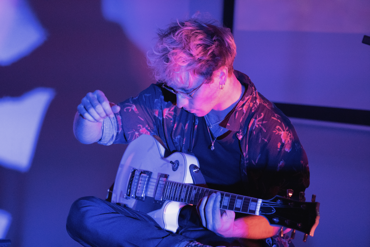 Musician with highlighted hair playing an electric guitar, intensely focused, with purple ambient lighting casting shadows in the background.