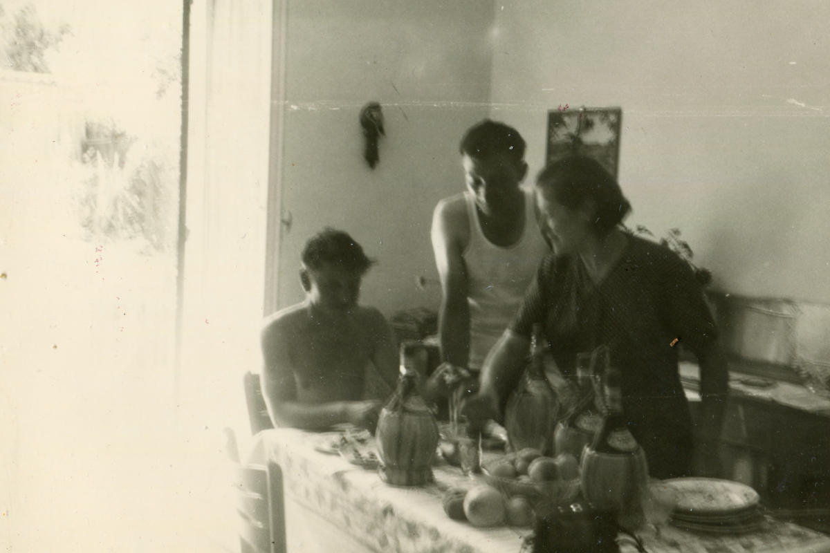 A vintage, black and white family scene. Three individuals are around a dining table adorned with several pitchers and fruit. A woman, standing, is actively engaging with a seated young boy, while a man in a tank top stands opposite the table, seemingly in mid-conversation or action. The image exudes a casual, intimate domestic setting, possibly capturing a moment of daily life. The sunlight streaming through a window on the left adds to the nostalgic quality of the photograph.