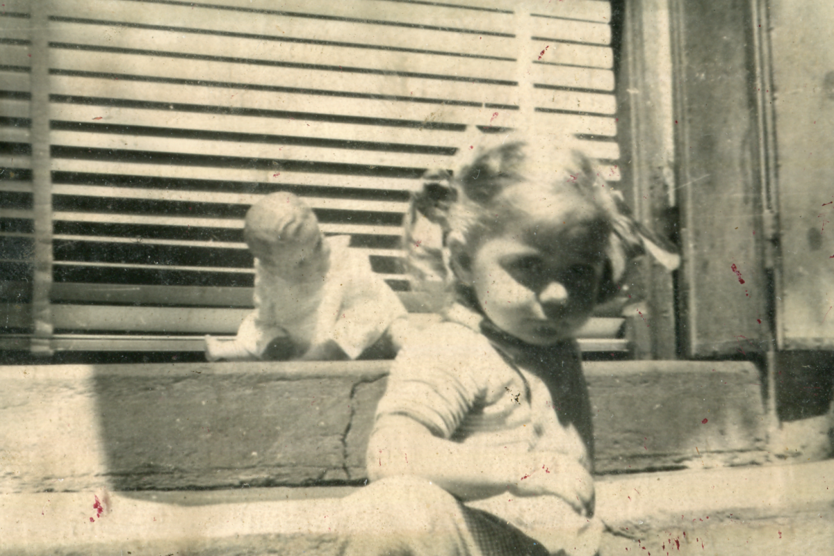An old, sepia-toned photograph showing two children sitting on a doorstep. The child in the foreground appears to be a young girl looking directly at the camera with a calm, perhaps curious expression. Her hair is styled in what seems like pigtails, and she is in a light-colored dress. The second child is in the background, with their back to the camera, focused on an activity out of view. They are both bathed in soft light, possibly indicating a sunny day. The texture and quality of the photo suggest it was taken several decades ago.