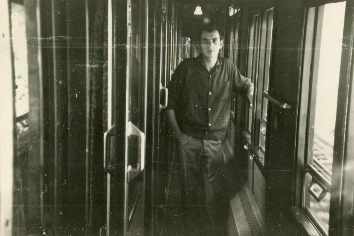 A vintage black-and-white photograph showing a young man standing in the corridor of a train. He is casually leaning on the frame of an open train door, looking directly into the camera with a calm and slightly inquisitive expression. The man is dressed in a dark, button-up shirt and trousers, with his hair neatly combed. The light coming from the windows of the train creates a high contrast in the corridor, casting deep shadows and bright highlights throughout the scene. The photograph has the characteristic imperfections of age, such as scratches and dust marks, which add to its historical ambiance.