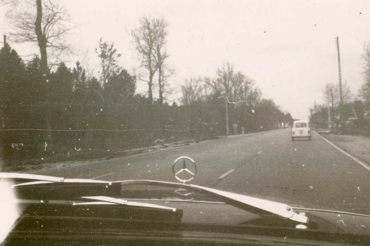 A black-and-white photograph taken from the perspective of a driver inside a vehicle, as indicated by the visible section of the dashboard and the iconic Mercedes-Benz emblem centered on the car's bonnet. The view is of a two-lane road stretching forward, lined with leafless trees suggesting a fall or winter season. There is a clear sky, and another vehicle is visible in the distance, driving in the same direction. The photograph has the hallmarks of an older film camera, with a grainy texture and some dust and scratches, which could suggest it's a vintage photo.
