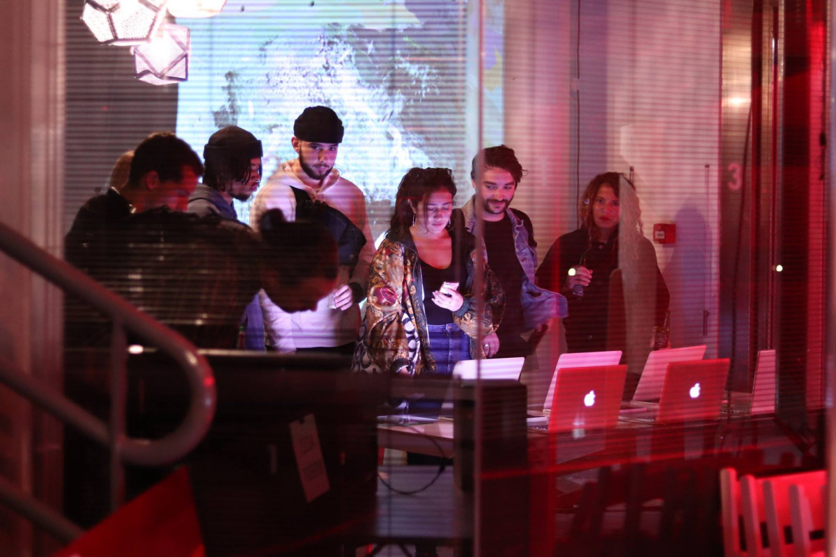 Group of people engaged with laptops, possibly participating in an interactive session, with glitch art projections illuminating the room in red hues at an art exhibition.