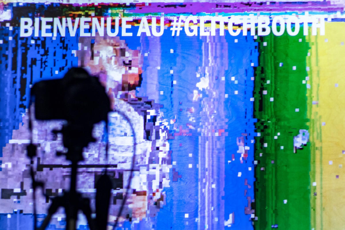 Silhouette of a camera tripod against a colorful glitch art background with text 'BIENVENUE AU GLITCHBOOTH' at an art exhibition.
