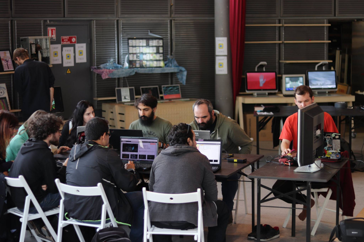 Workshop participants engaged in a glitch art workshop, seated around tables with laptops, electronic components, and monitors displaying digital art, in a spacious room with industrial aesthetics.