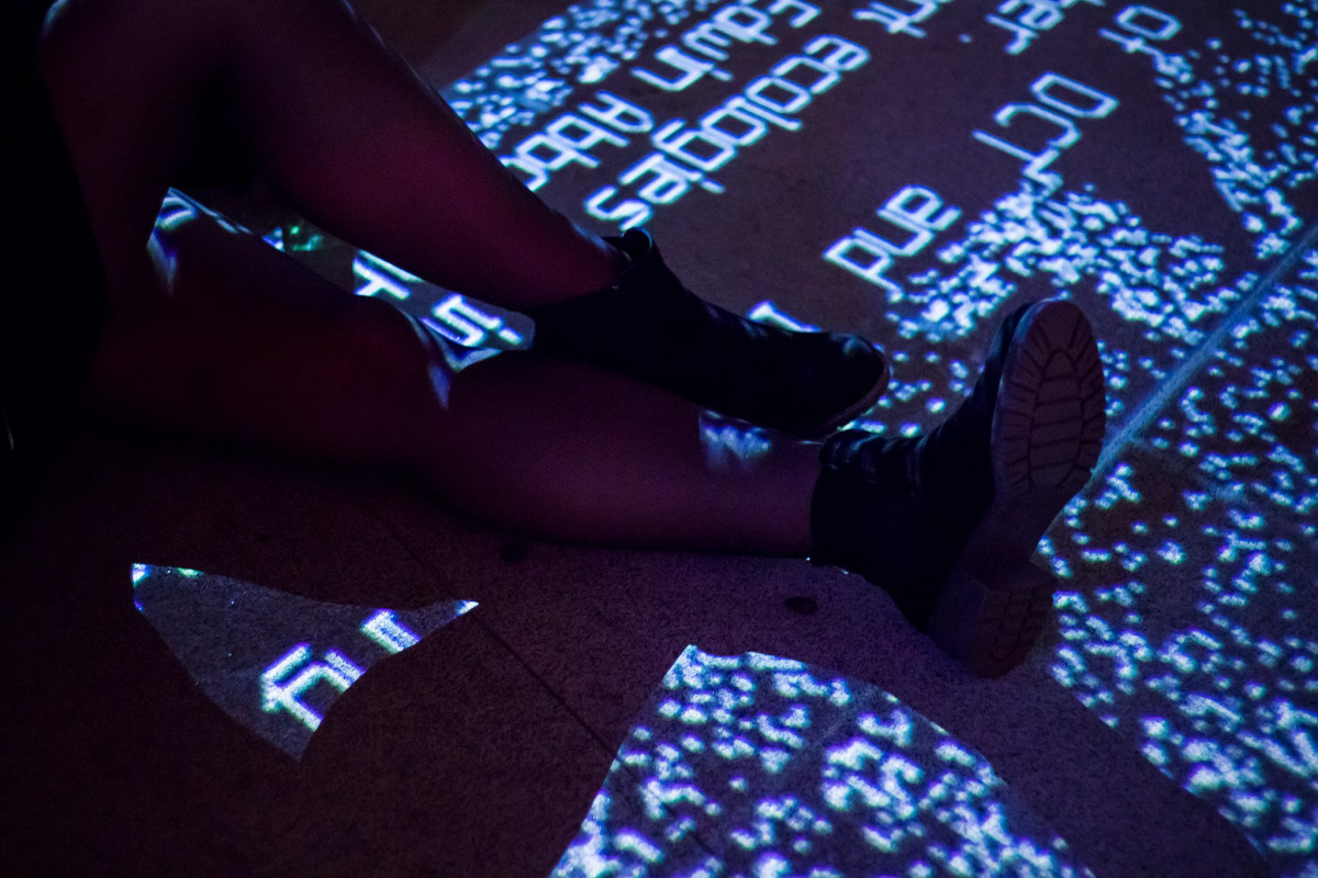 Close-up of legs with glitch art projections on them, creating a digital pattern overlay in a darkened room at a glitch art exhibition.