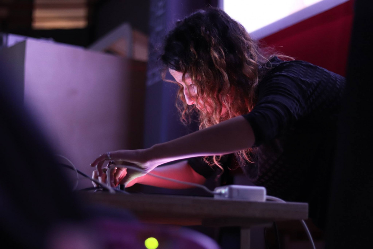 Sound artist performing at a gallery, intently manipulating electronic equipment under dim, ambient lighting.