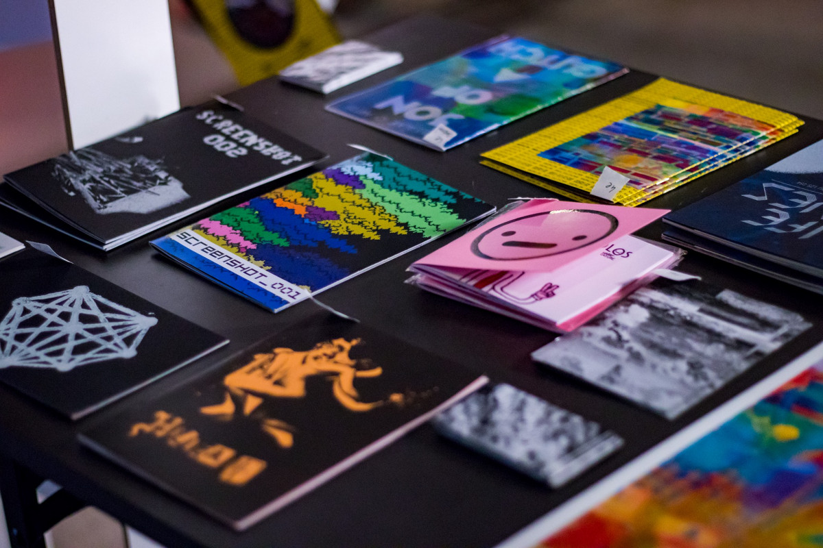 Table displaying an assortment of glitch art zines and printed materials with vibrant colors and various graphic designs at an art exhibition.