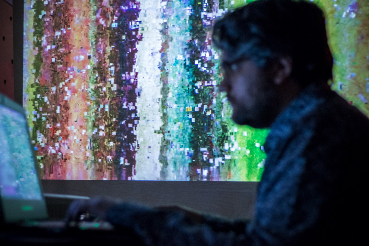 Artist creating glitch art, with a colorful digital mosaic projected in the background, illuminating the darkened space of the exhibition.