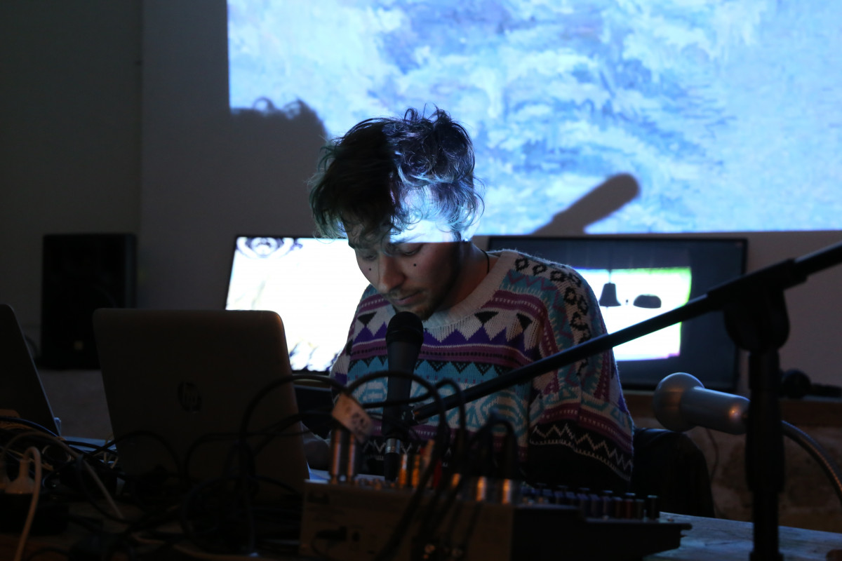 Focused individual with microphone and audio equipment, working on a laptop, with blue glitch art projections in the background during a live performance.