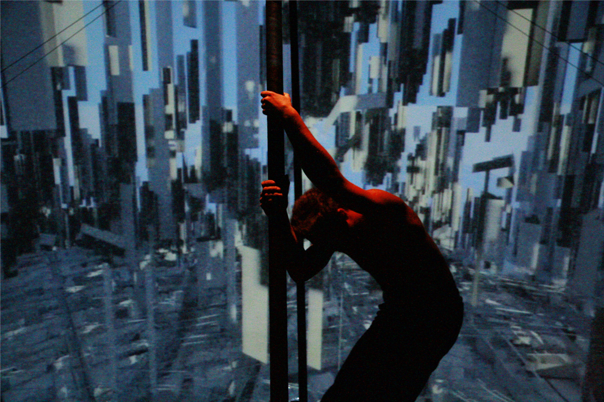 A Chinese pole performer in a dynamic, leaning pose with one arm outstretched, set against a projected backdrop of a digitally altered cityscape giving the illusion of a distorted urban environment.
