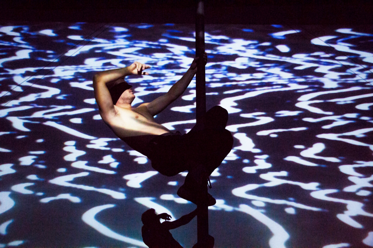 A shirtless Chinese pole artist performing an acrobatic move, cast in silhouette against a backdrop of abstract digital projections, creating a dynamic interplay of light and shadow.