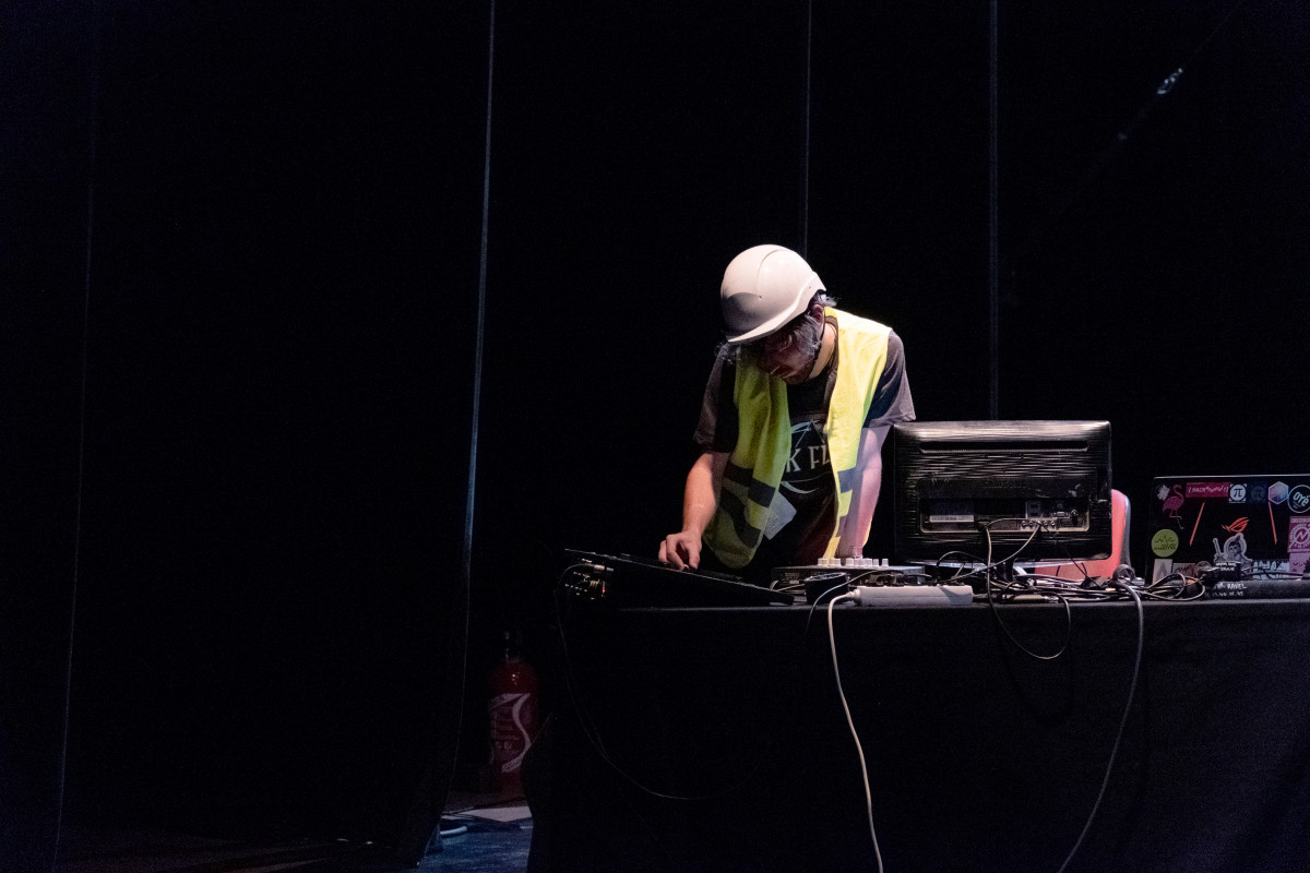 A person wearing a safety helmet and high-visibility vest focused on operating audio equipment, suggesting a performance art piece with a theme of construction or maintenance.