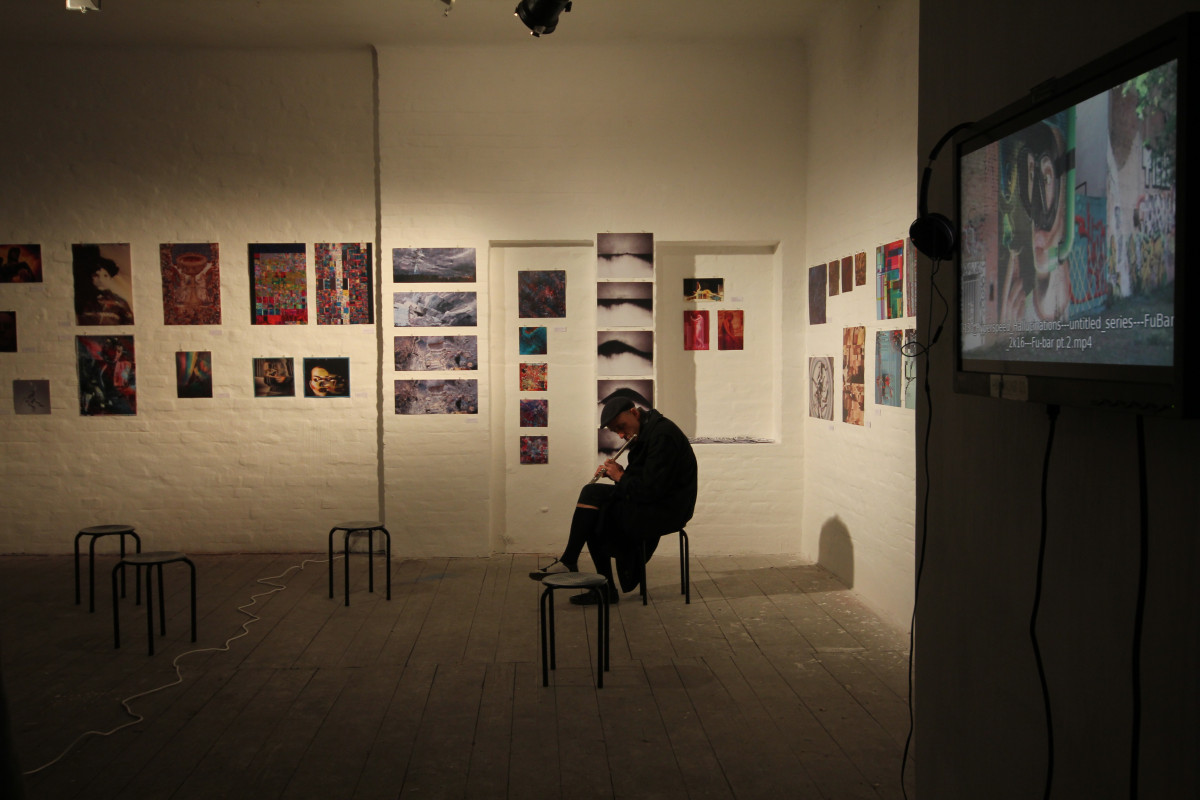 In a gallery with white brick walls adorned with various pieces of colorful and abstract art, a man sits on a black chair, deeply engrossed in playing a flute. To his right, a flat-screen monitor mounted on the wall displays a glitch art video, contributing to the creative and eclectic atmosphere of the room.