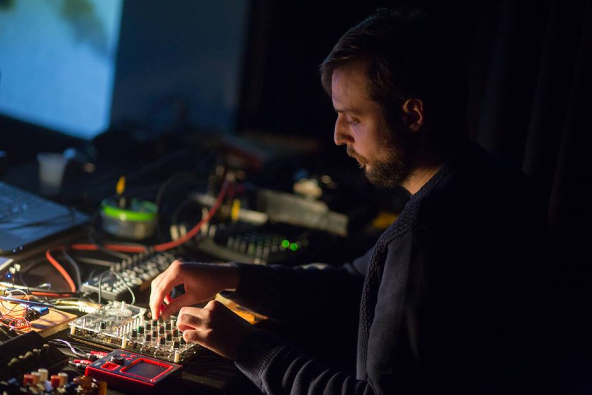 A focused sound artist engaged in a live performance, surrounded by an array of electronic devices and cables. The artist, a man with a beard and wearing a dark sweater, is intently manipulating a modular synthesizer and other equipment. The soft ambient lighting highlights his hands and the equipment, casting a warm glow amidst the dark surroundings.