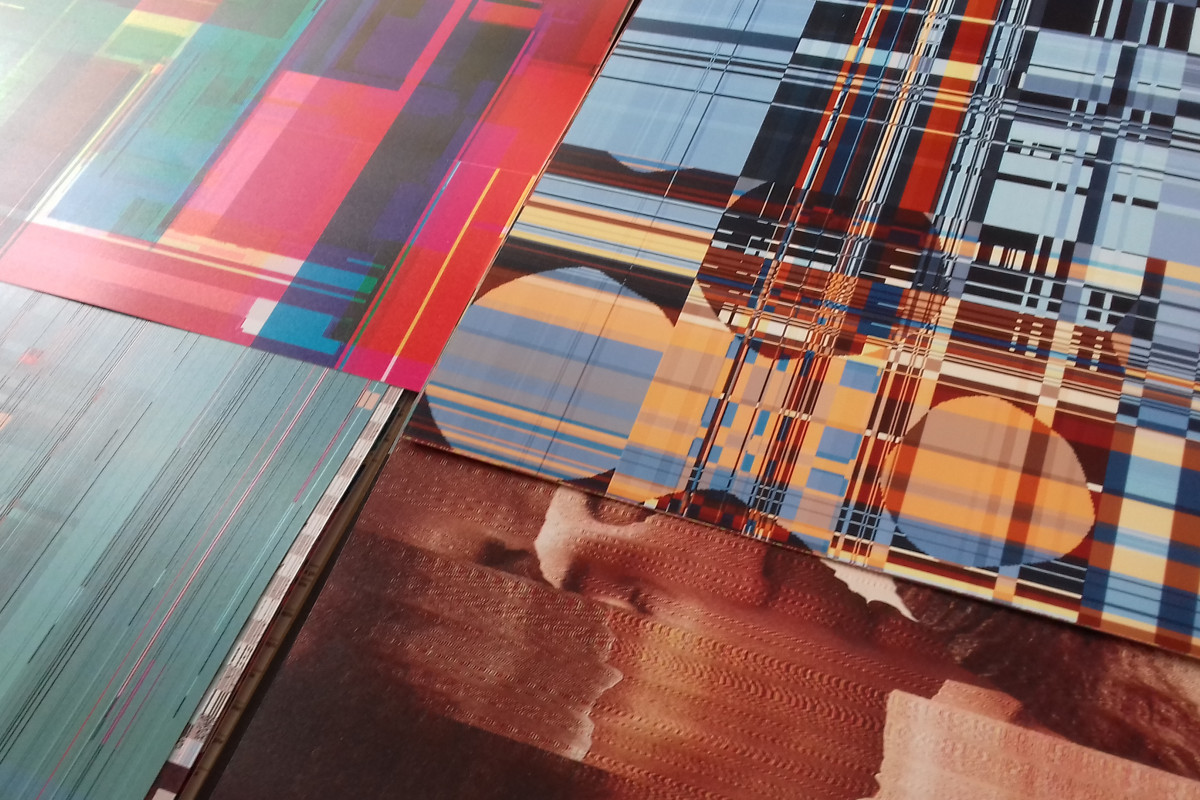 A close-up photo showing a collection of vibrant, abstract printed artworks. The prints feature a mix of digital glitch aesthetics with vivid colors and pixelated patterns. Foreground prints have streaks of cyan and magenta, while the background print combines grids and checkerboard patterns with orange and blue hues, all layered over a textured, earth-toned underlay.