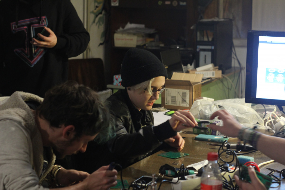 A group of individuals is engaged in an electronics workshop, focused on soldering and assembling components. Amidst a cluttered table full of wires, soldering irons, and electronic parts, a person with a black beanie and glasses is carefully handling a circuit board, while others are intently working on their own projects.