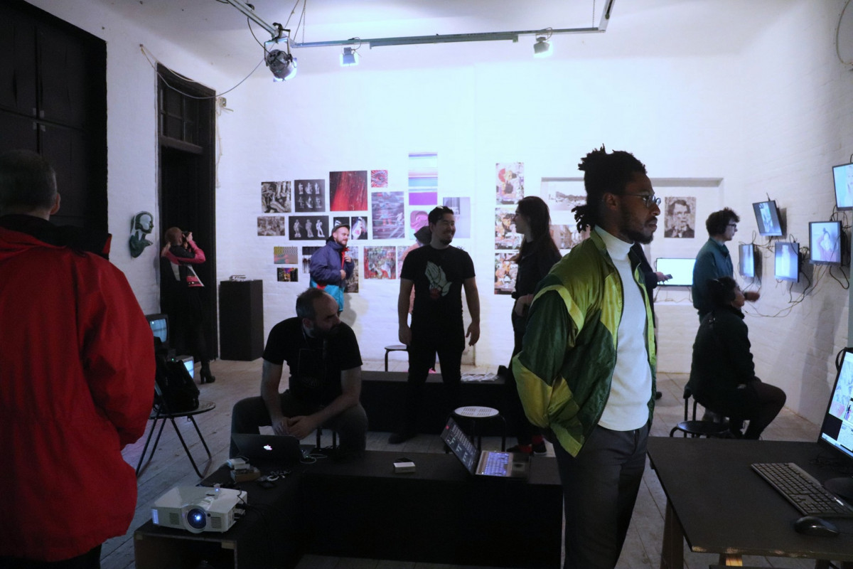 Visitors are scattered around a spacious gallery during an art exhibition. Artworks and digital screens adorn the white walls, illuminated by ambient lighting. In the foreground, a man in a red jacket observes the scene, while a person wearing headphones captures a moment with their camera. Various individuals are engaged with the exhibits, some in conversation while others are focused on the digital displays.