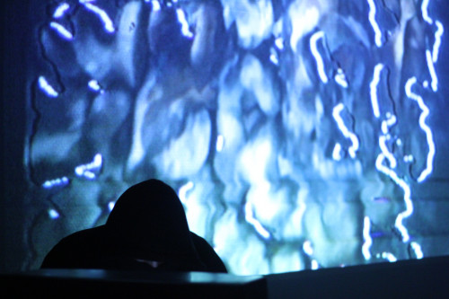 A visual artist performs in near darkness, their face concealed by a hooded sweatshirt. In the background, a vibrant display of analog glitch art with undulating patterns and electric blue streaks illuminates the scene.