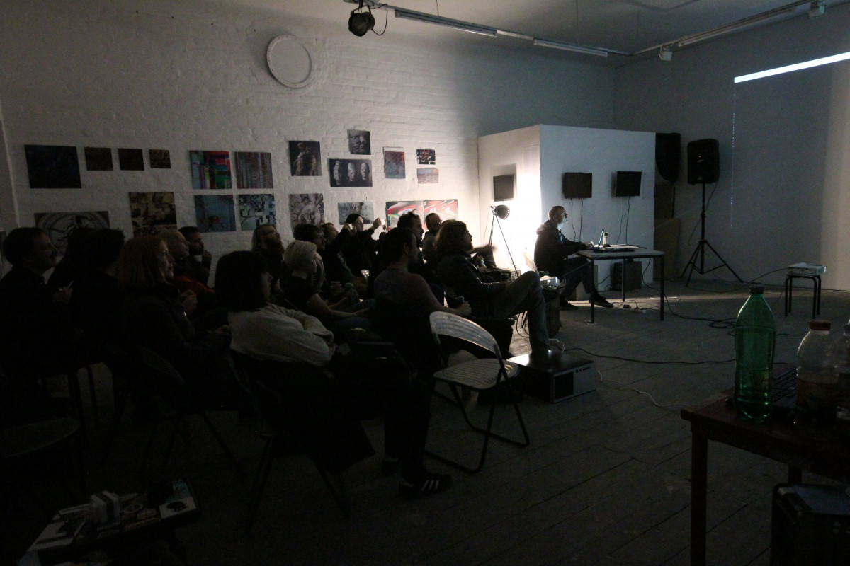 An image capturing a performance night at an art gallery, with a dark ambiance. The audience is seated in rows of chairs, their attention directed towards a screen emitting a soft glow on the right, where a person is seated at a desk with a laptop, possibly presenting. The left side of the room is adorned with an eclectic mix of artworks.