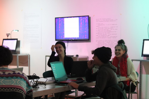 Group of people attending a glitch art workshop, with individuals smiling and interacting around laptops displaying colorful glitched images, in a room with text projections on the wall.