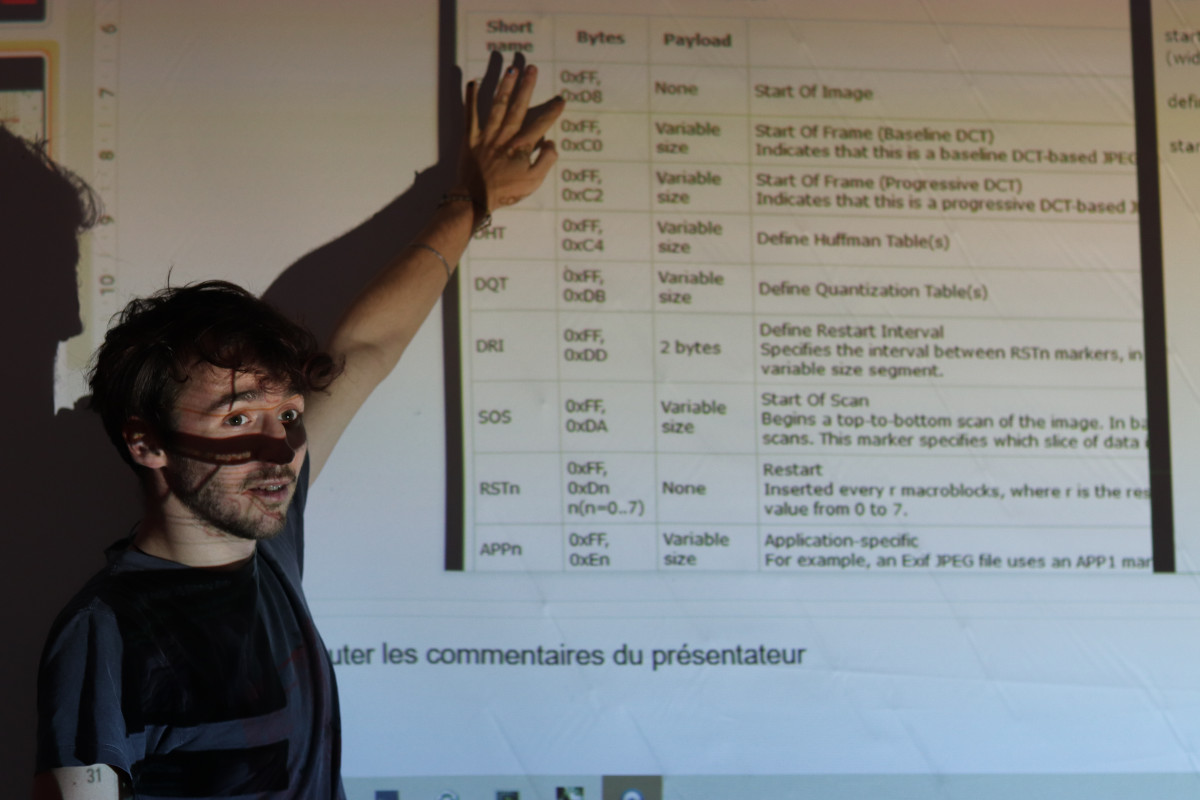 Presenter with a shadow of glitch art projections on his face, explaining JPEG structure on a screen during a workshop.