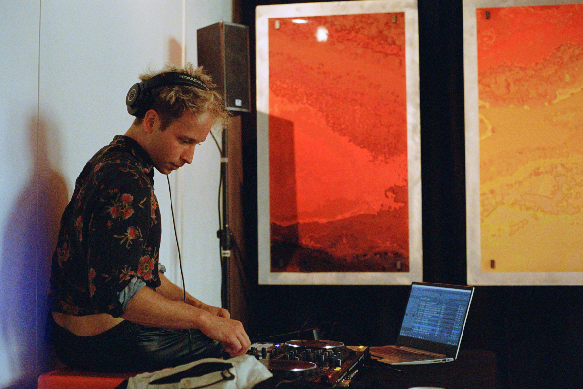 A DJ focused on his equipment, with headphones on, is playing music at an art exhibition opening. Behind him are large framed prints with abstract red and yellow designs.