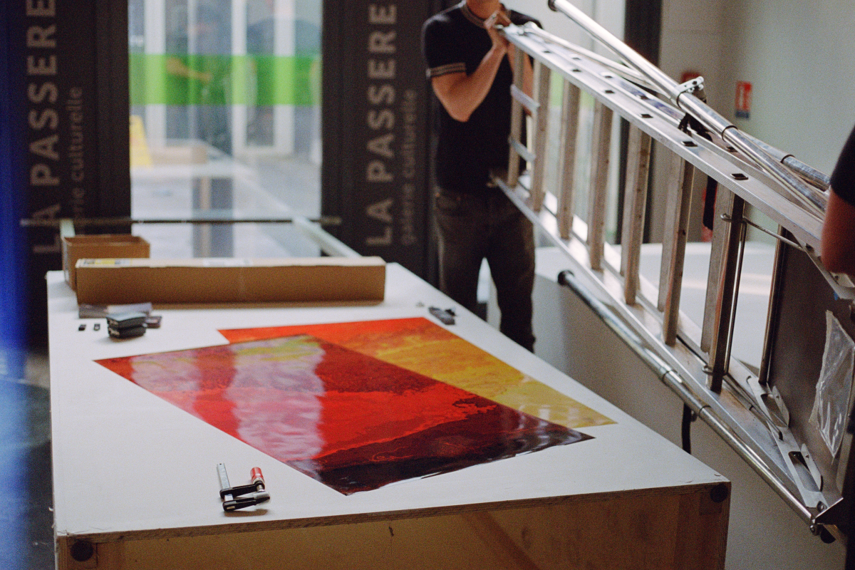 A person in a black shirt and jeans carries a ladder across a room where an art exhibition is being set up. On the table in the foreground, there's a large digital art print with bold red and yellow tones, alongside various setup tools like a staple gun and rolls of tape.