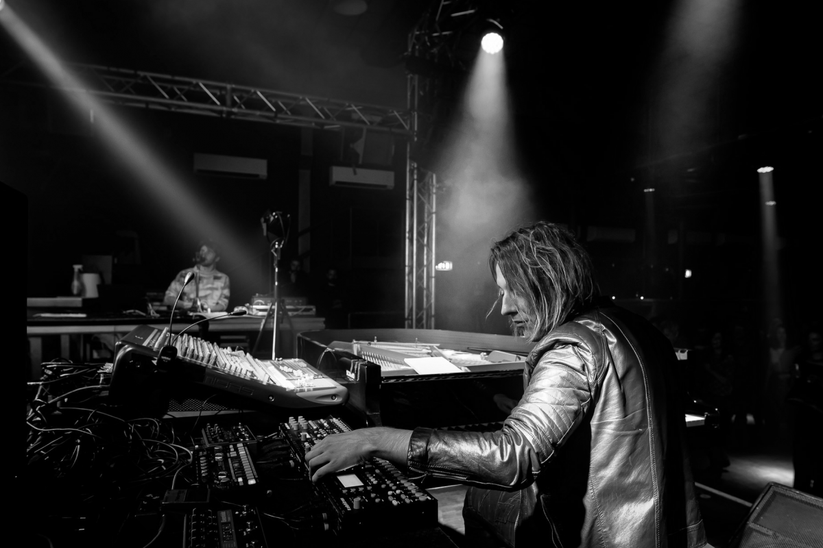 Black and white photo of a stage with a musician in a leather jacket operating electronic music equipment in the foreground and a VJ at the piano in the background, under dramatic stage lighting and haze.