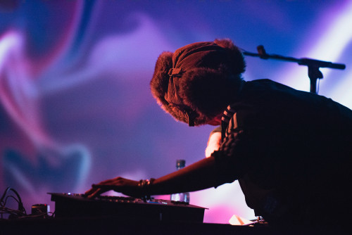 VJ wearing a winter hat adjusting equipment with abstract purple and blue visuals projected in the background, highlighting a creative performance atmosphere