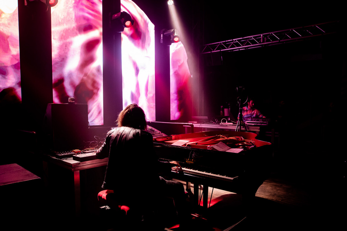 Pianist and VJ performing on stage with vibrant pink and white visuals on screens, amidst dramatic lighting, creating an artistic concert atmosphere.