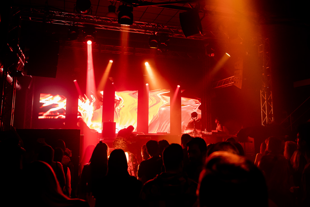 Concert scene with a pianist and a VJ performing on stage, red lights and abstract visuals on screens, and an engaged audience.